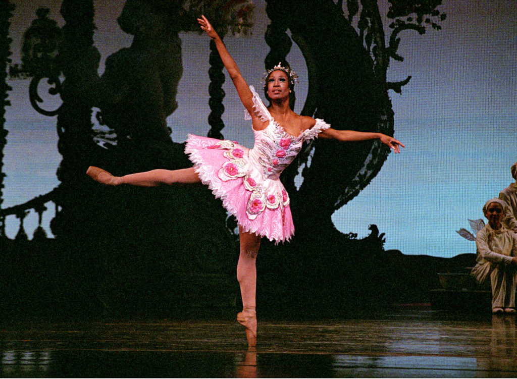 Lauren Anderson balances in an open arabesque en pointe, back arm raised on a diagonal. Her pointe shoes and tights are dyed brown to match her skin tone. She wears a pink and white tutu and a tiara.