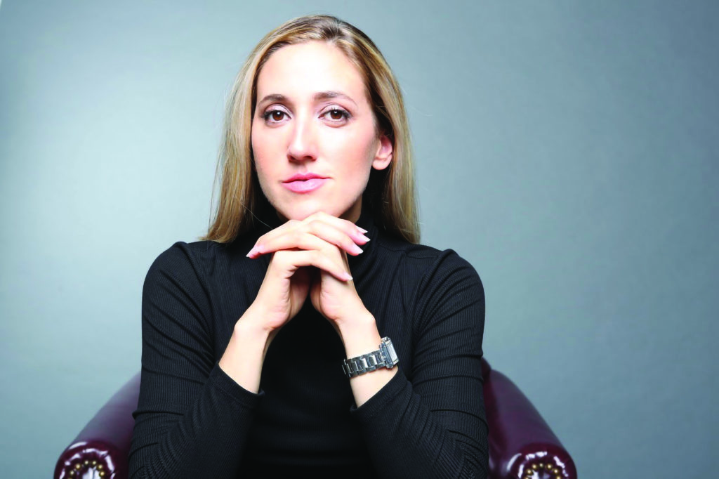 Melissa Barak sits with her hands clasped below her chin, gazing seriously at the camera. Her dirty blonde hair is loose and disappears behind her shoulders. She wears a black, long-sleeved shirt, and is seated in a burgundy armchair.