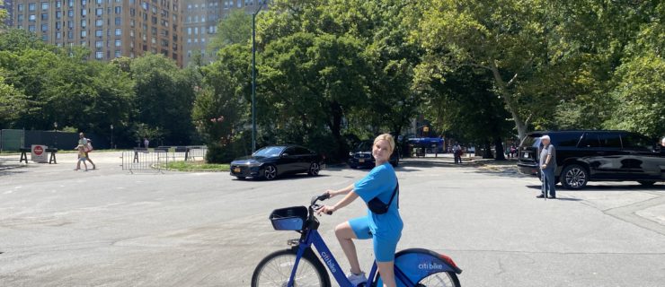 HALEY HILTON is a fair-skinned blonde woman wearing a blue shirt and blue bike shorts, riding a blue Citi Bike on a sunny day in Central Park.