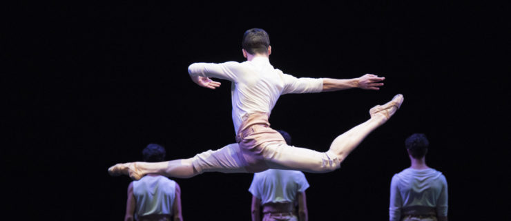 In the foreground, a male dancer leaps with his back leg in attitude, torso twisting to face upstage, arms in opposition to legs. Another dancer sits on the floor, looking up at him. In the shadowy upstage, three dancers stand with their backs to the audience. All wear white shirts, khaki shorts, and ballet slippers.