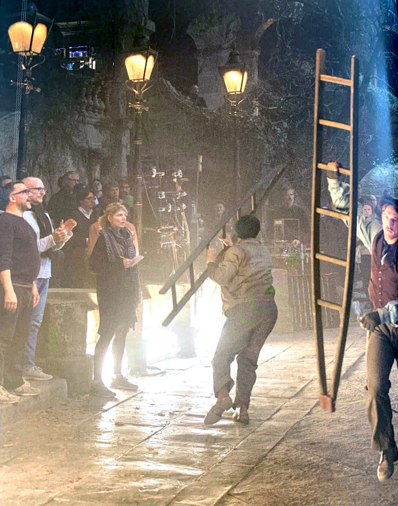Tara Nicole Hughes stands wearing layered street clothes, eyes on performers with a notebook in hand. In the foreground, performers in period wear rush about with ladders, lit by street lanterns. A mass of crew members are in the background, carefully off-camera.