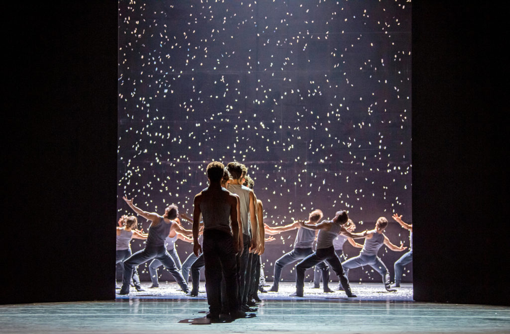 Upstage, a black scrim opens partway to reveal dancers in deep fourth position pliés, arms stretched wide and chests uplifted to what looks like falling snow. A line of dancers downstage faces them, as though waiting in line to join them.