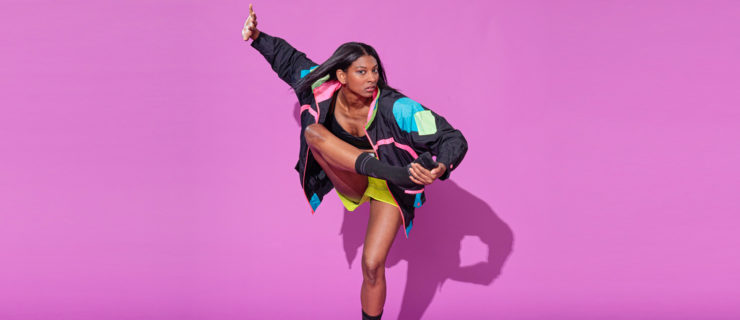 Micaela Taylor poses on a bright purple backdrop. She balances on one leg as she raises the other toward her chest, holding her foot with her opposite hand. Her free arm rises to the side of her head, as though pushing away the space. She wears dark socks, shorts, and a black jacket with geometric blocks of bright color.