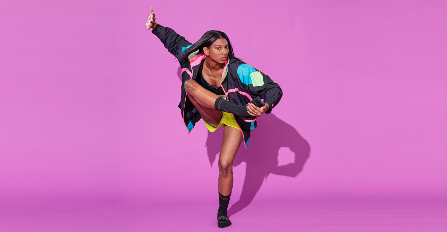 Micaela Taylor poses on a bright purple backdrop. She balances on one leg as she raises the other toward her chest, holding her foot with her opposite hand. Her free arm rises to the side of her head, as though pushing away the space. She wears dark socks, shorts, and a black jacket with geometric blocks of bright color.