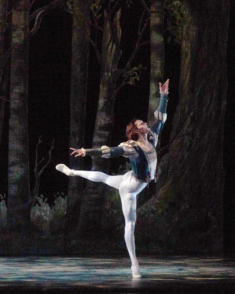 Daniel Camargo balances in back attitude croisé, chin upraised, his upstage arm extended high overhead, the other to the side. He wears a costume of white tights and ballet shoes, and a green, long-sleeved tunic with puffy white accents at the elbows and shoulders. The backdrop is of a shadowy wood.