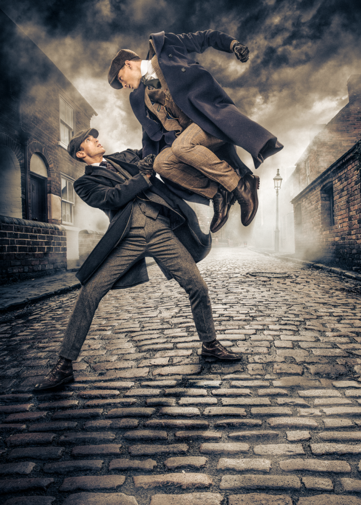 In a promotional image for "Peaky Blinders: The Redemption of Thomas Shelby," a man flies above the shoulders of his fellow, fist pulled back as though about to throw a punch. They wear three-piece suits, jackets, and caps appropriate for early-20th-century England. A murky cobbled street invaded by smoke and fog provides the backdrop.