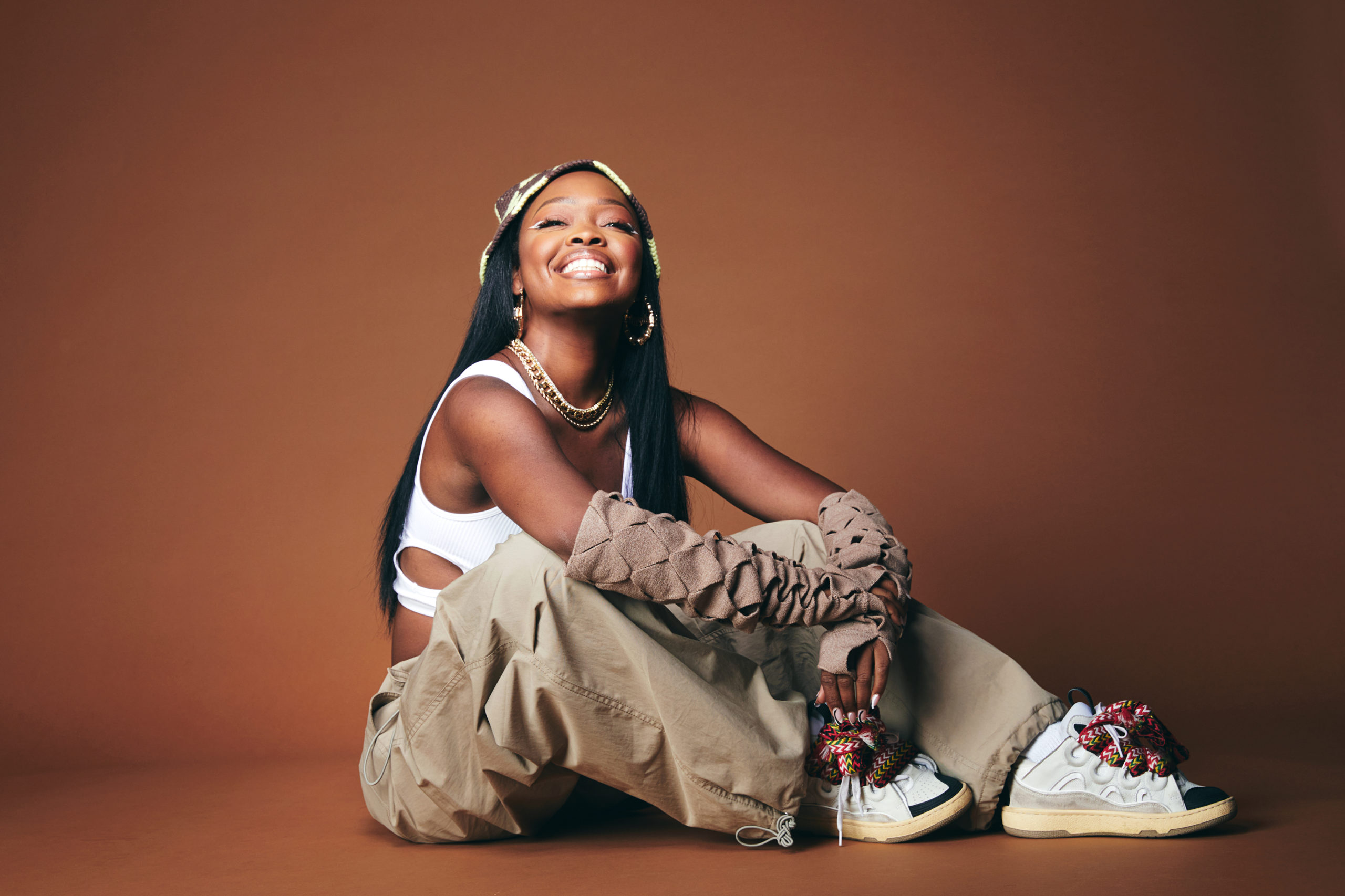 Charm La'Donna sits against a dark orange backdrop, smiling hugely at the camera. Her elbows are draped over her knees, one wrist clasping the other. She wears chunky sneakers, baggy beige pants with matching arm warmers, and a white crop top. Her black hair is straight and falls to her waist. She wears hoop earrings and a flat beaded necklace.
