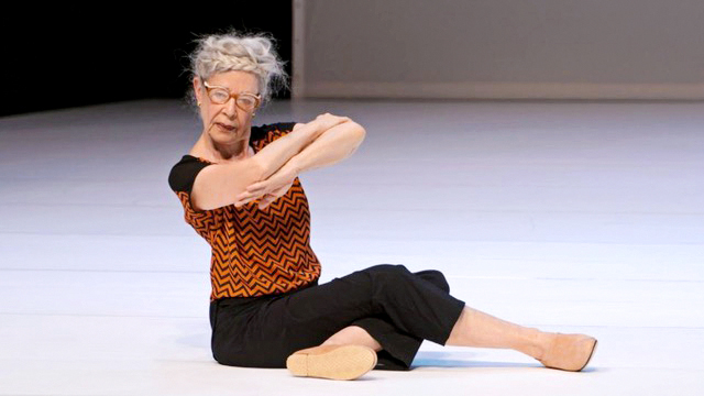 Deborah Hay sits onstage with her legs crossed in front of her. Her forearms are stacked, and she tilts them slightly downstage as she looks out to the audience. Her silver hair is braided in a crown around her head; she wears glasses and an orange and black patterned top.