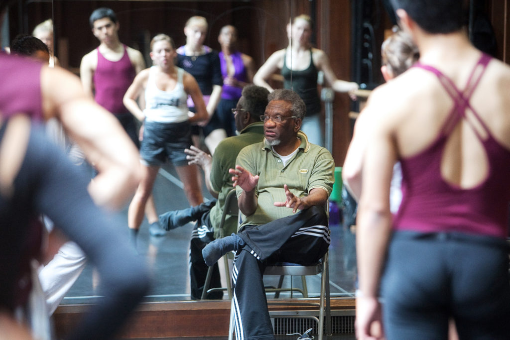 Man sitting in chair talking to group of dancers in studio