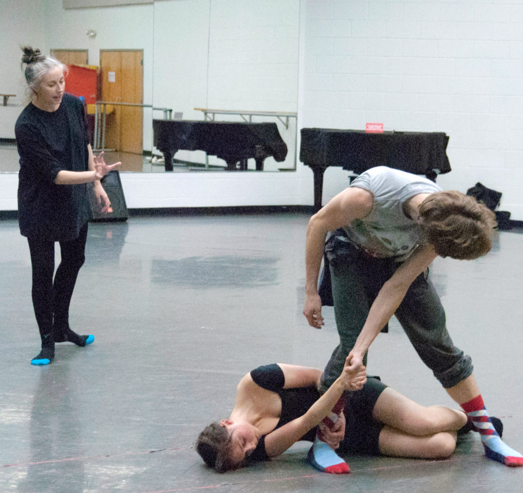 In the foreground, a dancer lies on her side, curled around the legs of another. The standing dancer bends forward, clasping the prone dancer's hand. Danielle Rowe observes from behind them, gesturing as she gives direction.