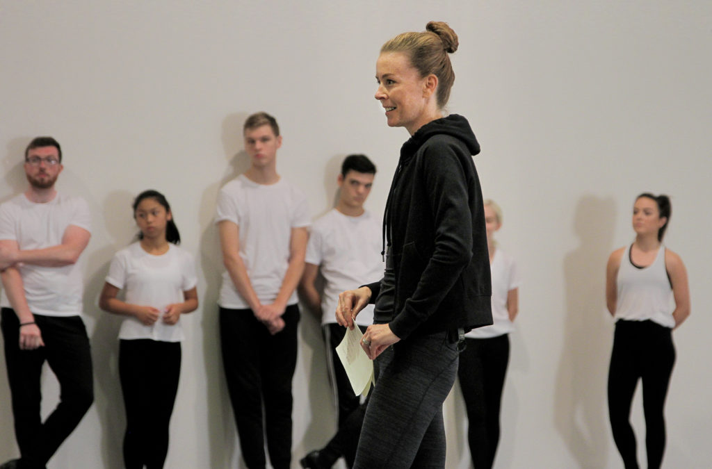 Jean Butler is seen in profile, smiling intently while holding a folded sheet of written notes.  In the blur behind her are a number of young dancers, all dressed in white shirts and black pants, leaning against a white wall, listening.