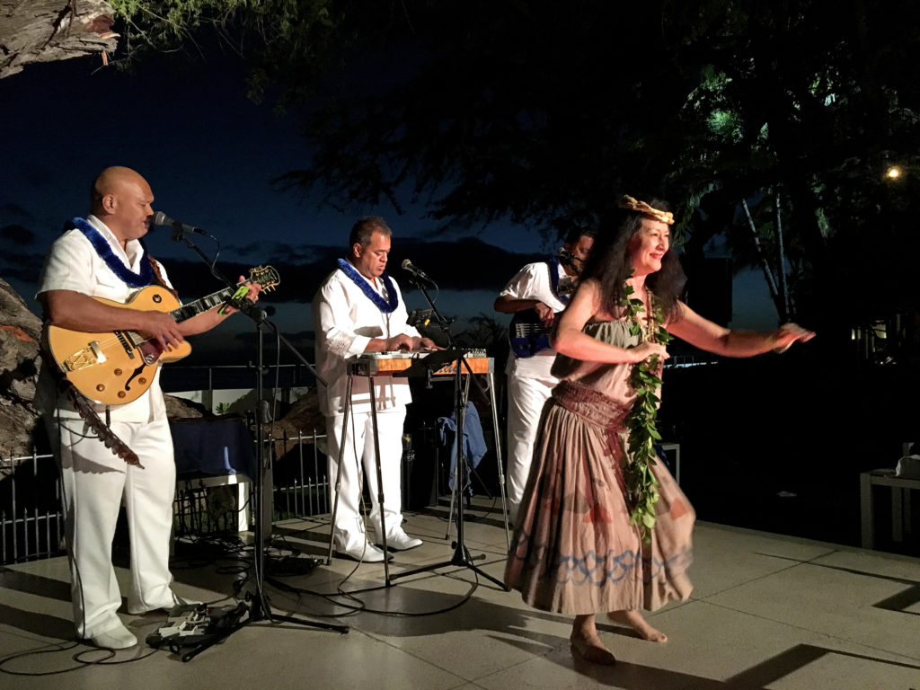 Kanoe Miller smiles, an arm extended toward her audience caught in motion blur as she transfers her weight on bent knees. Three musicians in white line the stage behind her. Trees, water, and islands are dimly visible in the darkness outside.