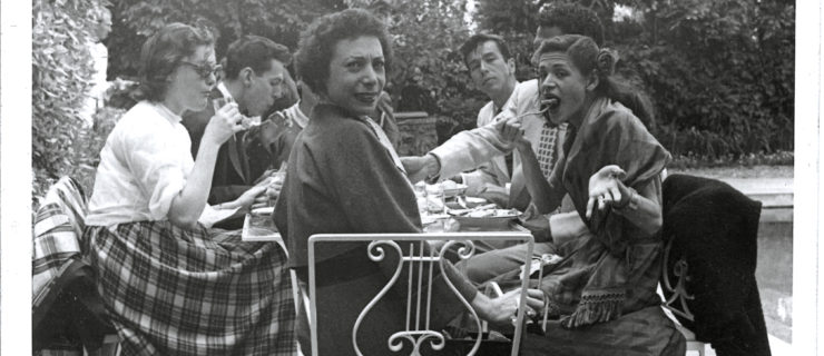 A black and white snapshot shows seven people sitting at a table outside next to a pool. One woman turns to look over her shoulder at the camera, while another mischievously scoops a large bite towards her mouth with a fork. The rest of the table continues eating and chatting behind them, unperturbed.