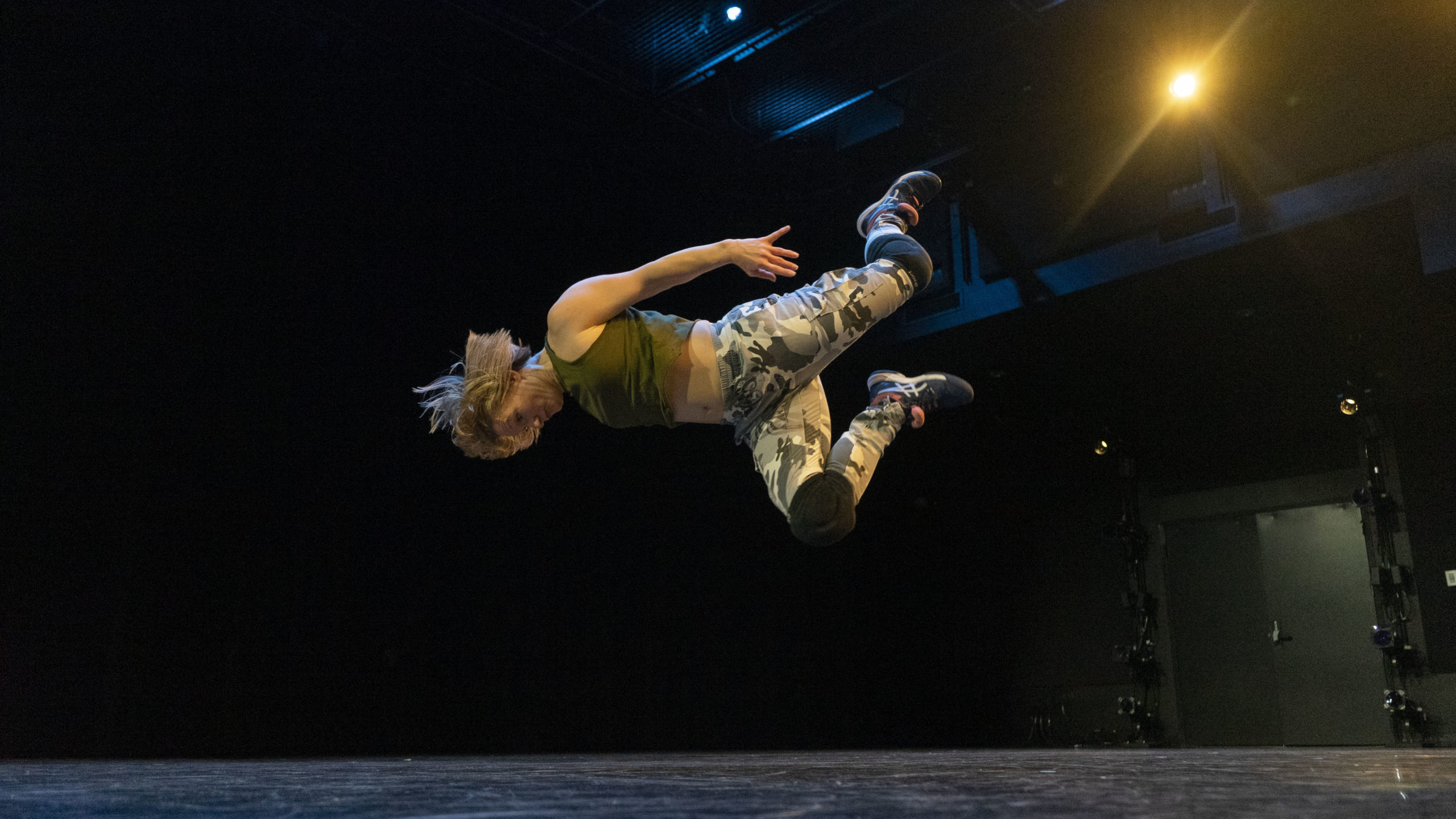 A dancer is caught mid-air, body parallel to the ground. Their arms are thrust behind them, knees kicked up. It seems impossible that they could catch themself before crashing to the ground.