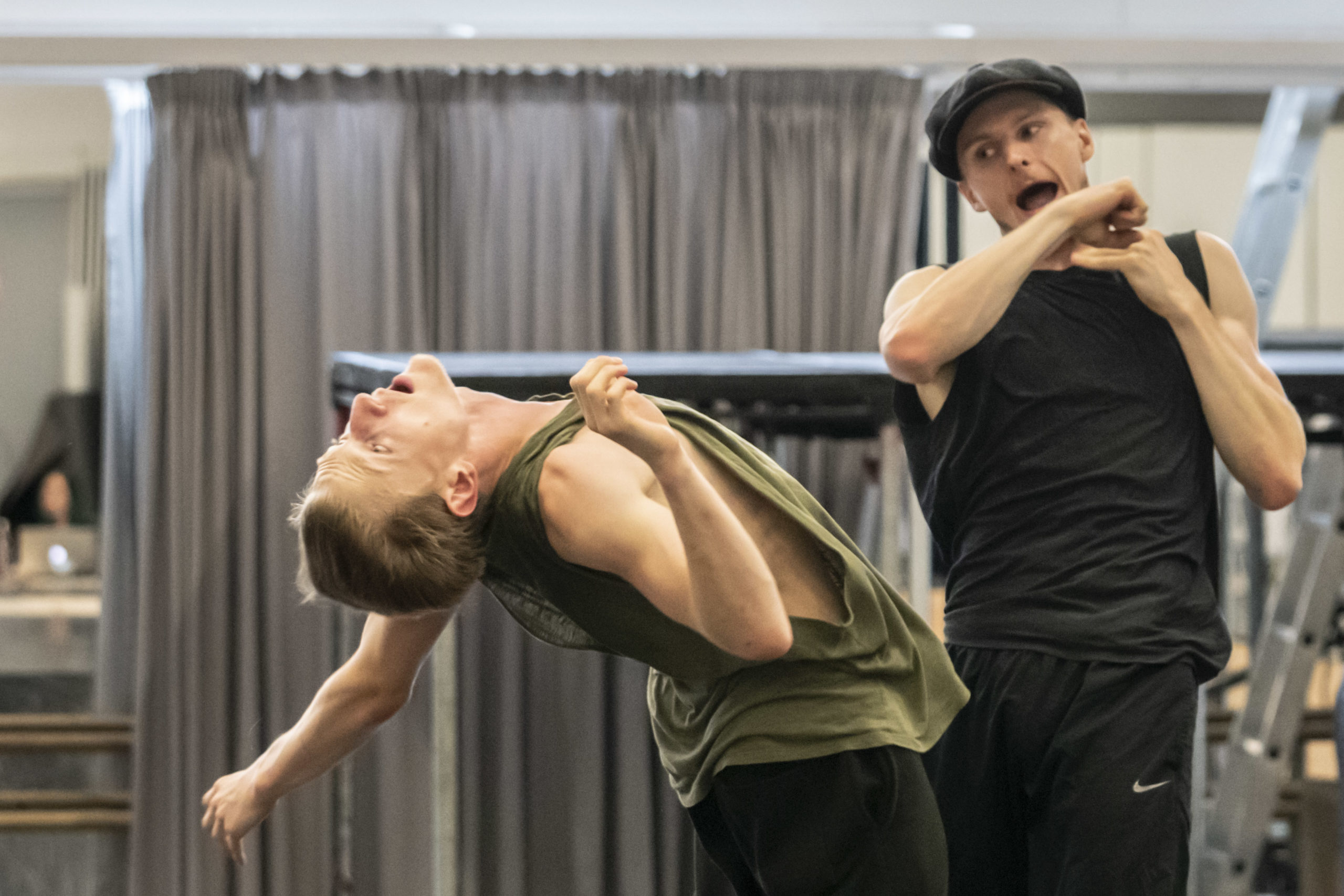In a rehearsal studio, a male dancer arches back, arms akimbo like he is flailing after being hit. Behind him, another dancer finishes the swing of his clasped fists, mouth open wide like he is yelling, a cap worn on his head an addition to the more standard rehearsal clothes.
