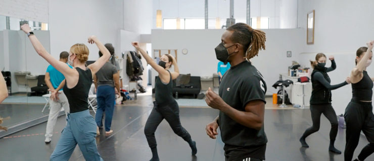 Kirven Douthit-Boyd is surrounded by dancers in movement throughout a studio with white walls and black floors.