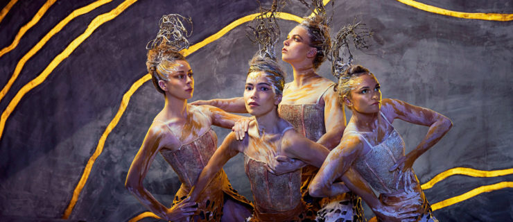 Four women cluster together, leaning in disparate directions with their ribcages with hands planted on hips or resting on each other's shoulders. Pale grey paint splatters across skin and hair, from which nests of silver wire rise. Gold veins cut through the dark backdrop.