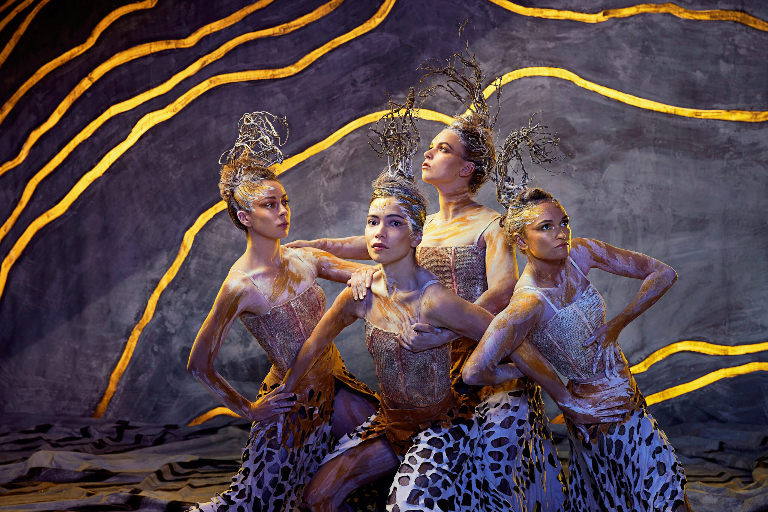 Four women cluster together, leaning in disparate directions with their ribcages with hands planted on hips or resting on each other's shoulders. Pale grey paint splatters across skin and hair, from which nests of silver wire rise. Gold veins cut through the dark backdrop.