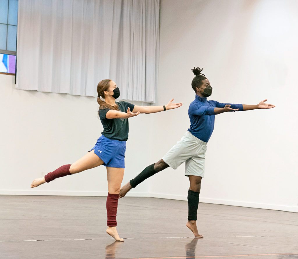 Dancers Karlie Budge, who is wearing a black shirt, and Brandon Randolph, who is wearing a blue shirt, are both standing on one leg with their arms outstretched before them.