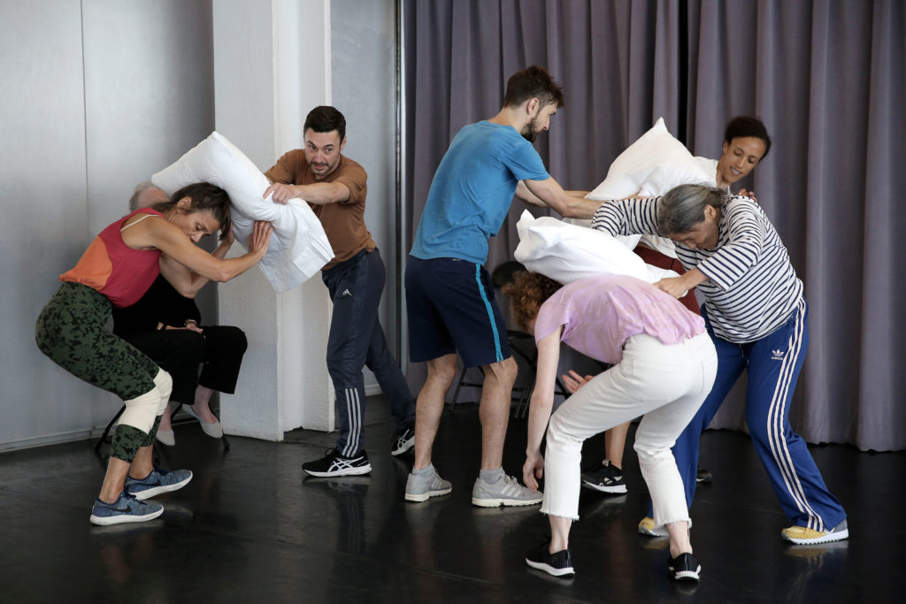 Six dancers of varying ages, genders, and ethnicities are in the midst of what seems to be a pillow fight. All wear variations of athletic clothes and sneakers.