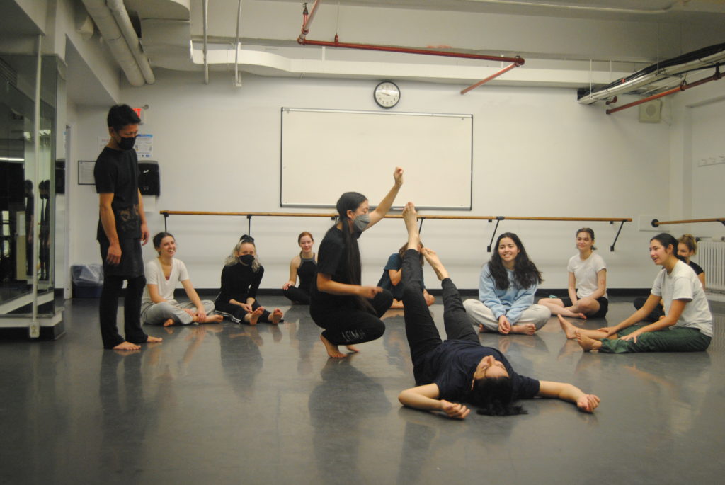 A dance student lies on her back with her feet suspended in the air as the instructor directs her and the class looks on.