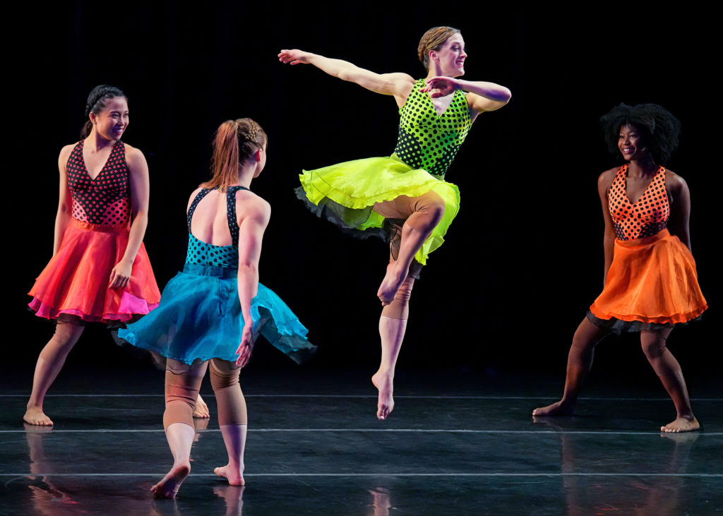 A barefoot dancer in a lime green dress that flares around her knees jumps in parallel passé, opposite arm flung straight behind her. A trio of dancers in matching dresses in different colors surround her, smiling, as they shift through plié.