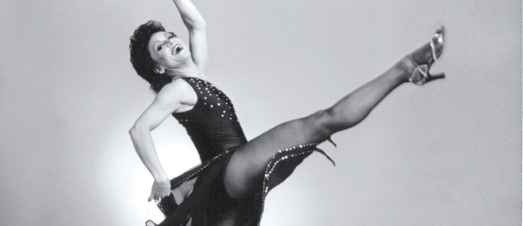 In a black and white image, Chita Rivera grins broadly as she moves through a shallow layout. Her back arm is curved overhead, the other flick her long skirt back. She wears a fitted bodice, flowing skirt, dark tights, and heels.