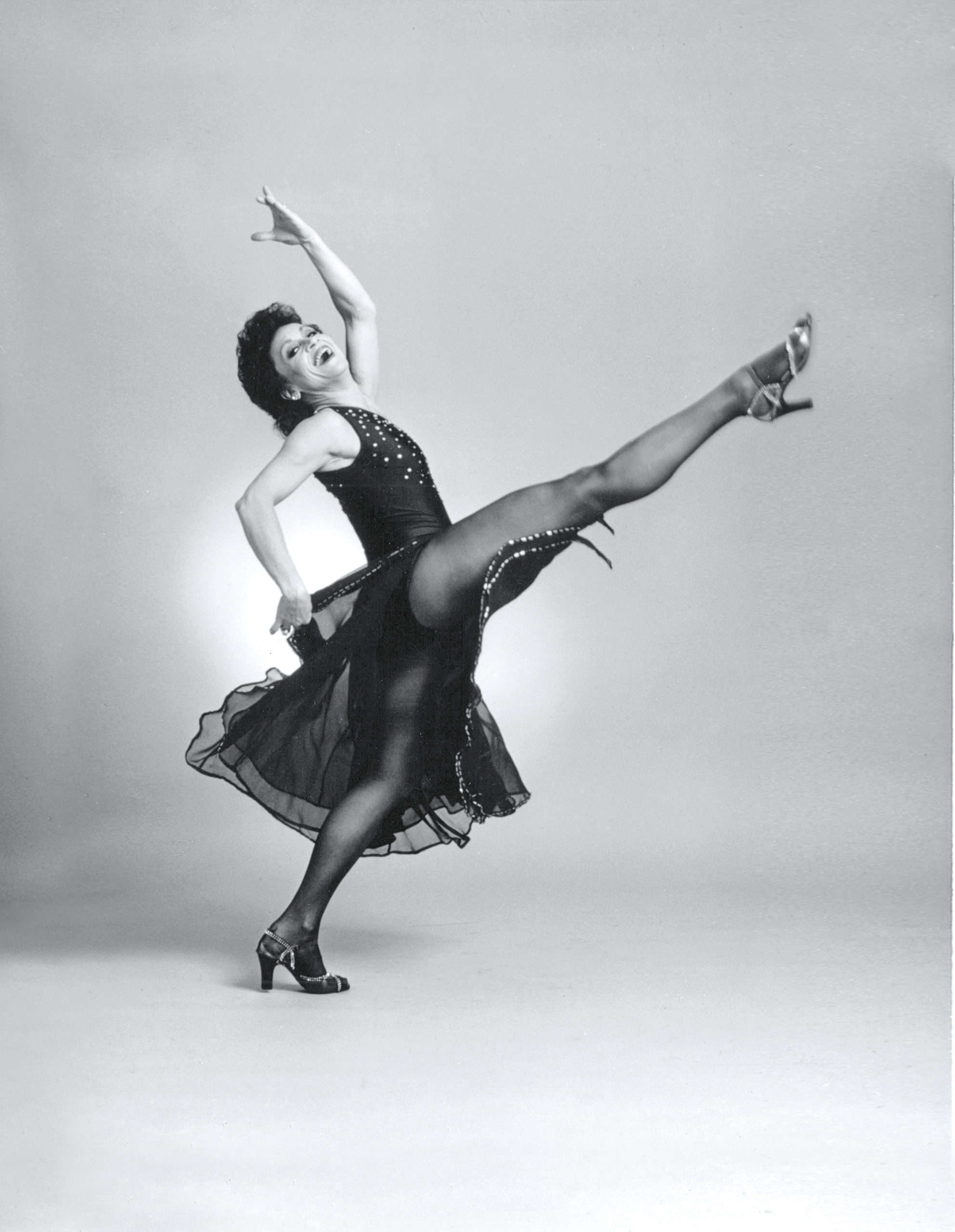 In a black and white image, Chita Rivera grins broadly as she moves through a shallow layout. Her back arm is curved overhead, the other flick her long skirt back. She wears a fitted bodice, flowing skirt, dark tights, and heels.