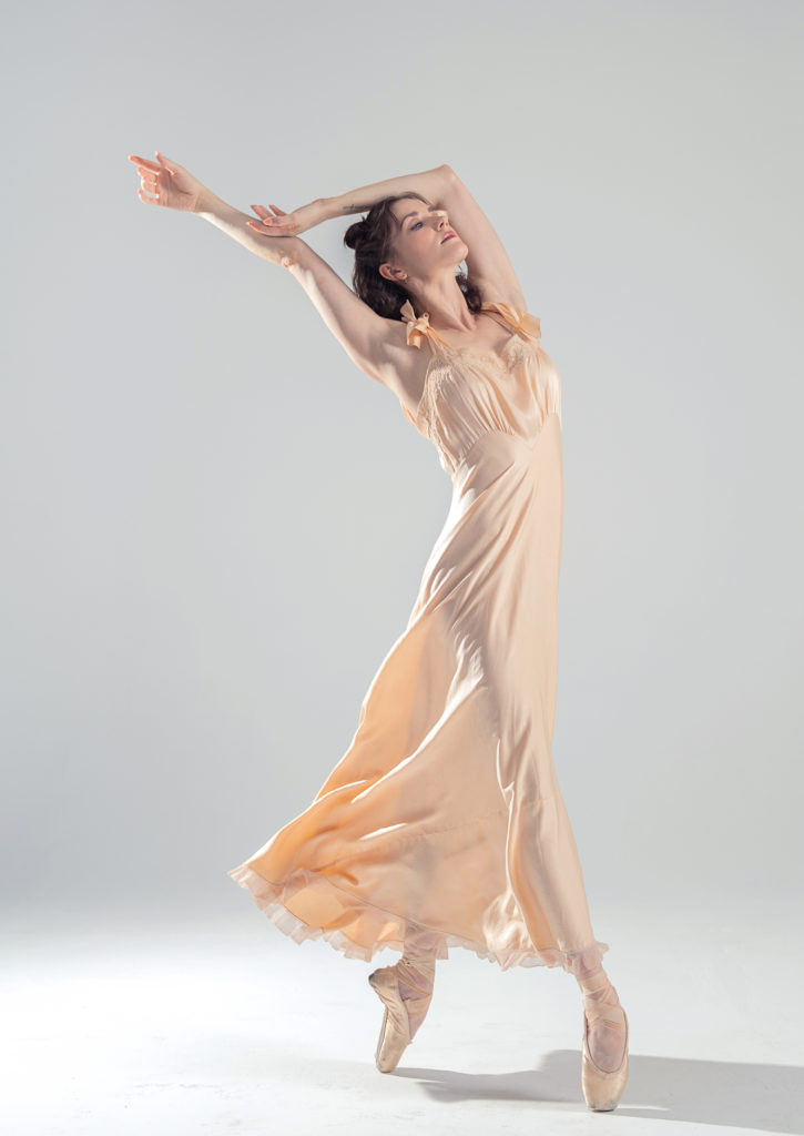 A dancer in a peach satin dress stands on pointe one foot crossed in front of the other arms up and behind her looking up to the right.