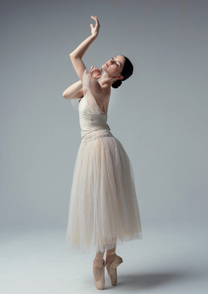 A dancer in a long white tutu stands on pointe, legs together one arm raised and the other bent in front of her as she gazes upward