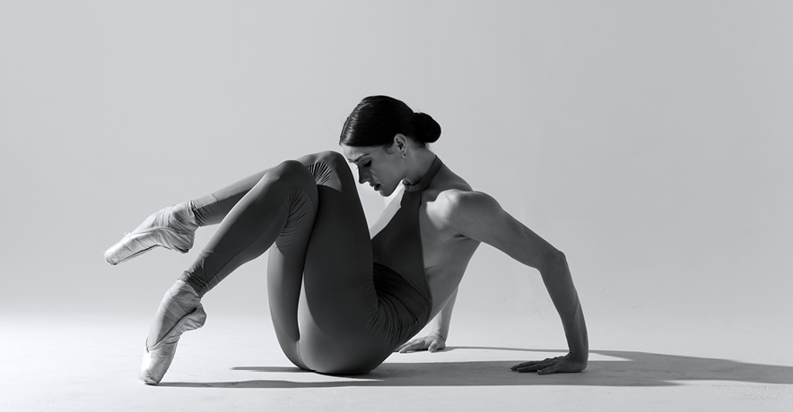 Black and white image of a dancer sitting with knees crossed and lifted up to her forehead, in a unitard and pointe shoes
