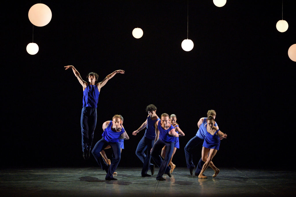 A dancer in dark trousers and a bright blue shirt jumps straight in the air, arms flying into a V beside his ears. A cluster of a half dozen other dancers are to the right, leaning forward with hands behind their backs as they pause in coupe back, knees bent.