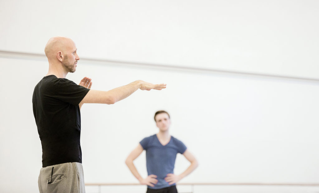 Wayne McGregor is caught in profile in a white-walled studio. He gestures with one arm forward, a flattening gesture, as he speaks to the room. A dancer is visible from the waist up, out of focus, listening with hands on hips.