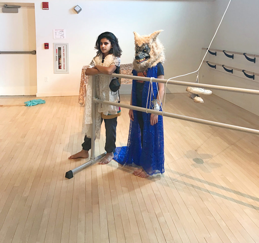 Two individuals stand at a barre set at an angle into the studio. One, in layers of rehearsal-wear, leans her elbows upon it and gazes distrustfully at the camera. The other, beside her, wears a flowing blue lace gown and a wolf head. Rope is tied to the barre and is rigged overhead out of sight; a pair of white sneakers dangles by its laces.