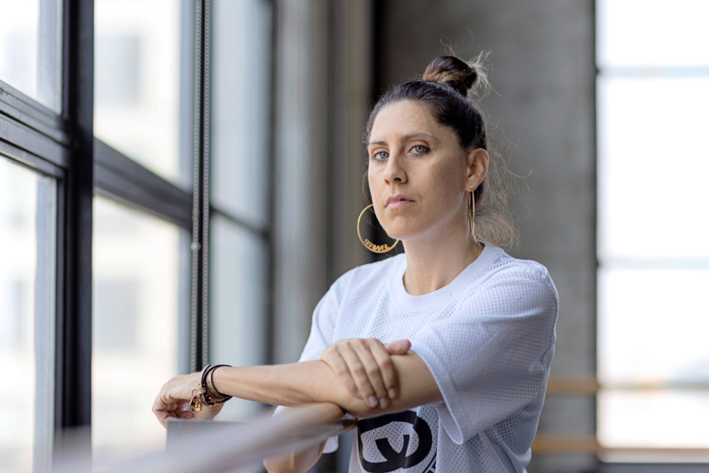 Jennifer Weber leans her elbows on a barre, posture upright as she gazes seriously at the camera. Her brown hair is pulled into a poofy bun on the top of her head. She wears gold hoop earrings and a white graphic tee.