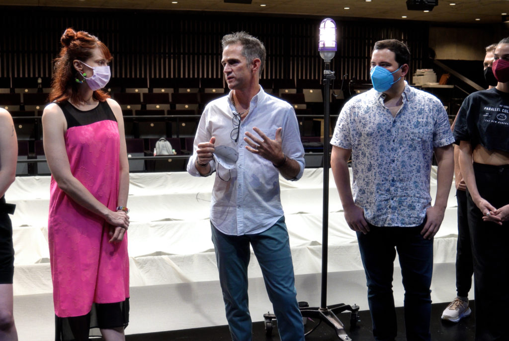 Andy Blankenbuehler stands beside a ghost light in a largely empty theater as he speaks to a gathered group. Those in view wear face masks.