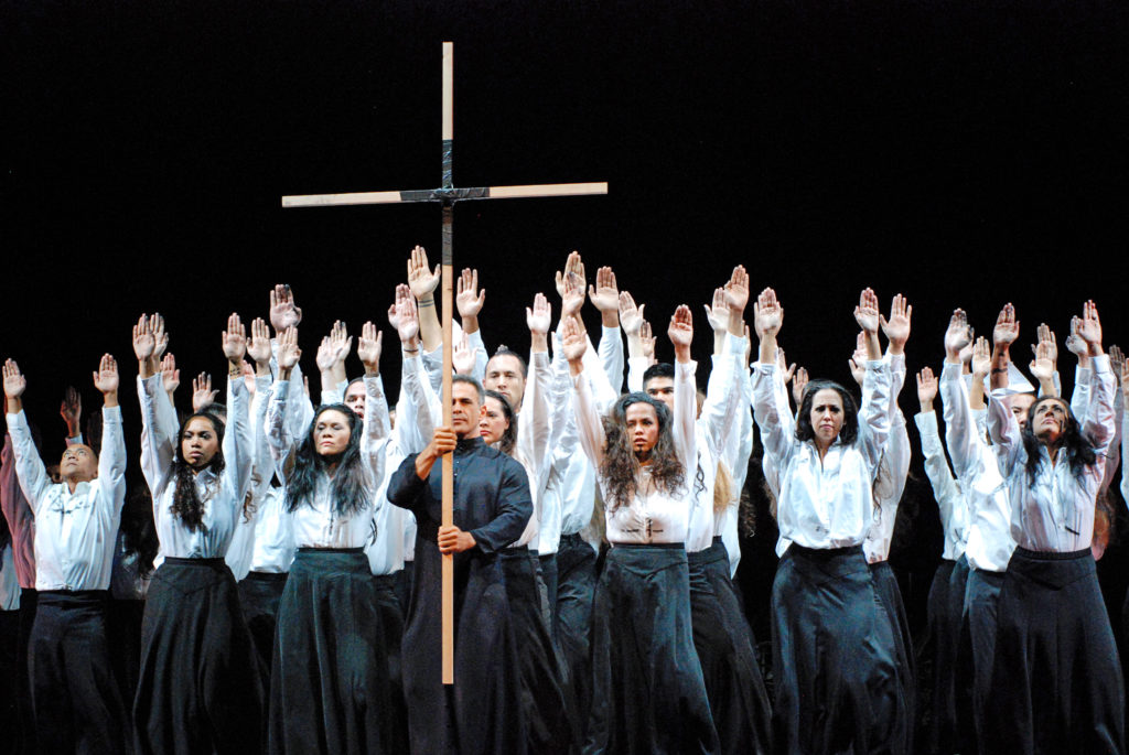 large group of dancers wearing white shirts and black skirts, man carrying a cross 