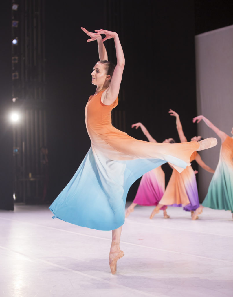 Cecilia Iliesiu turns en pointe in attitude back, arms in high fifth, focus out to the audience. The perspective is from the wings. She wears a long, flowing dress of orange, blue, and white.