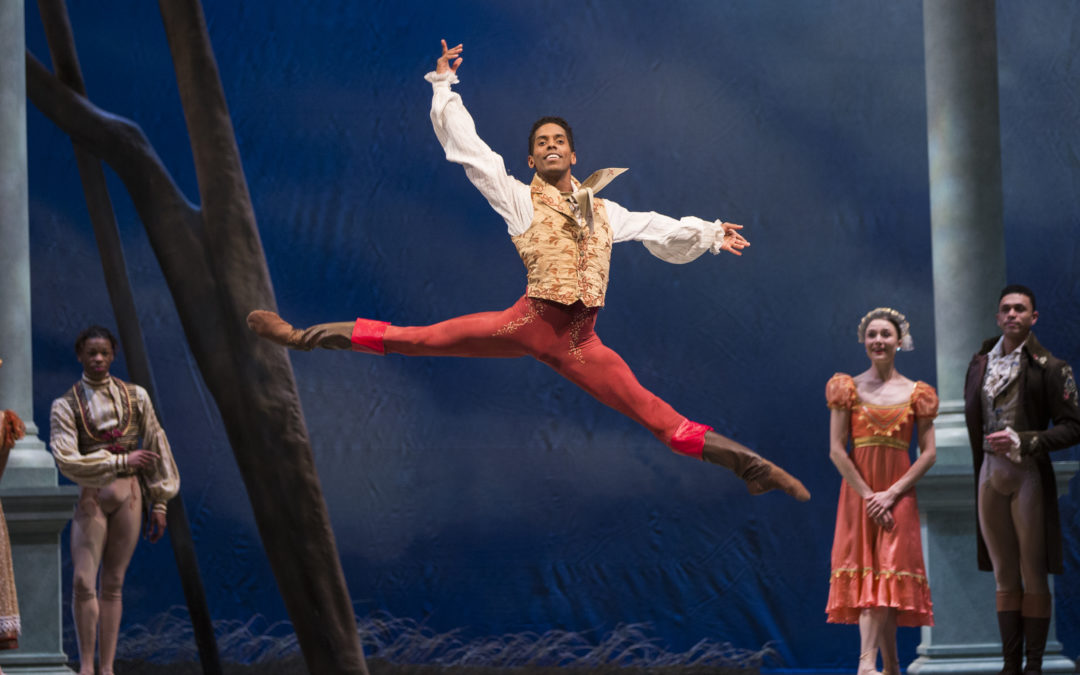 Jonathan Batista leaps, back leg and arm raised high as he lifts his chin to the audience. Costumed dancers watch from the sides and upstage. He is costumed in brown boots, patterned red tights, and a fitted gold vest over an old-fashioned white shirt with puffy sleeves.