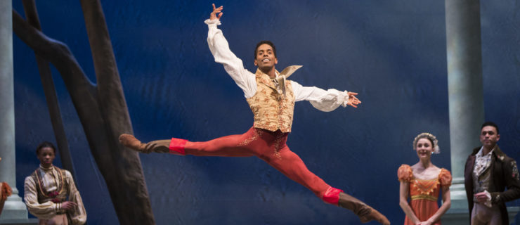 Jonathan Batista leaps, back leg and arm raised high as he lifts his chin to the audience. Costumed dancers watch from the sides and upstage. He is costumed in brown boots, patterned red tights, and a fitted gold vest over an old-fashioned white shirt with puffy sleeves.