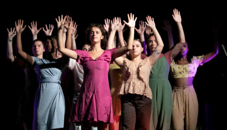 A tight cluster of teenaged dancers in muted blouses and dresses stand with their hands raised by their heads under a harsh light.