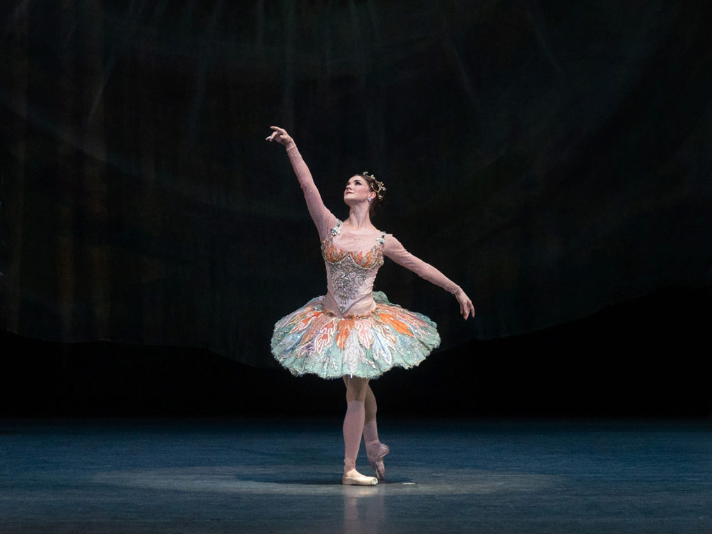 In performance, a dancer in an orange and green tutu stands on one leg the other pointed behind her and one arm raised, the other to the side at hip level