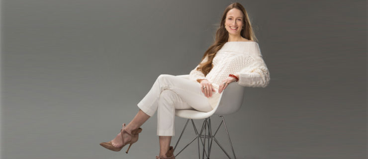 Julie Kent sits in a white chair turned to the side. She smiles broadly at the camera, her long brown hair falling over her shoulders to her waist. She wears a white, off-the-shoulder sweater, white trousers, and brown heels.