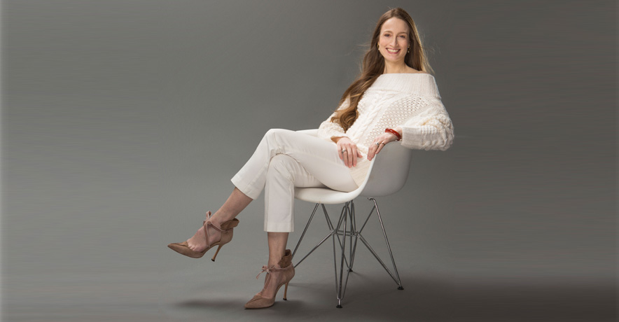 Julie Kent sits in a white chair turned to the side. She smiles broadly at the camera, her long brown hair falling over her shoulders to her waist. She wears a white, off-the-shoulder sweater, white trousers, and brown heels.