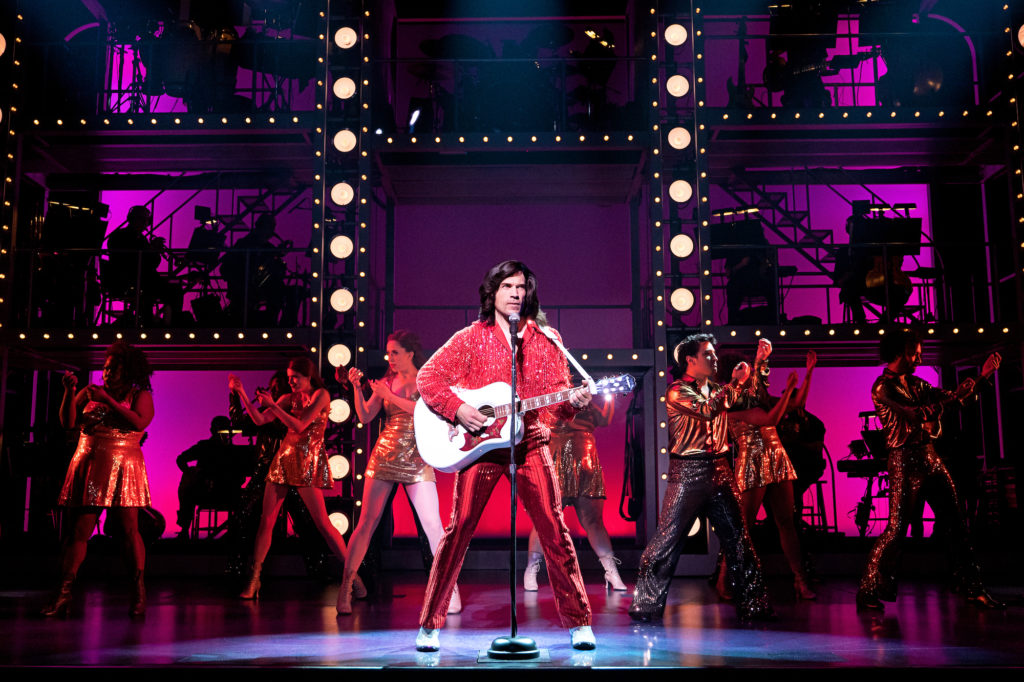 Will Swenson stands onstage in front of a microphone in a wide stance as he strums a white guitar with red accents. His costume is red and shiny. In the background, a pyramid of male and female dancers in shiny gold costumes gesture in mirrored unison.