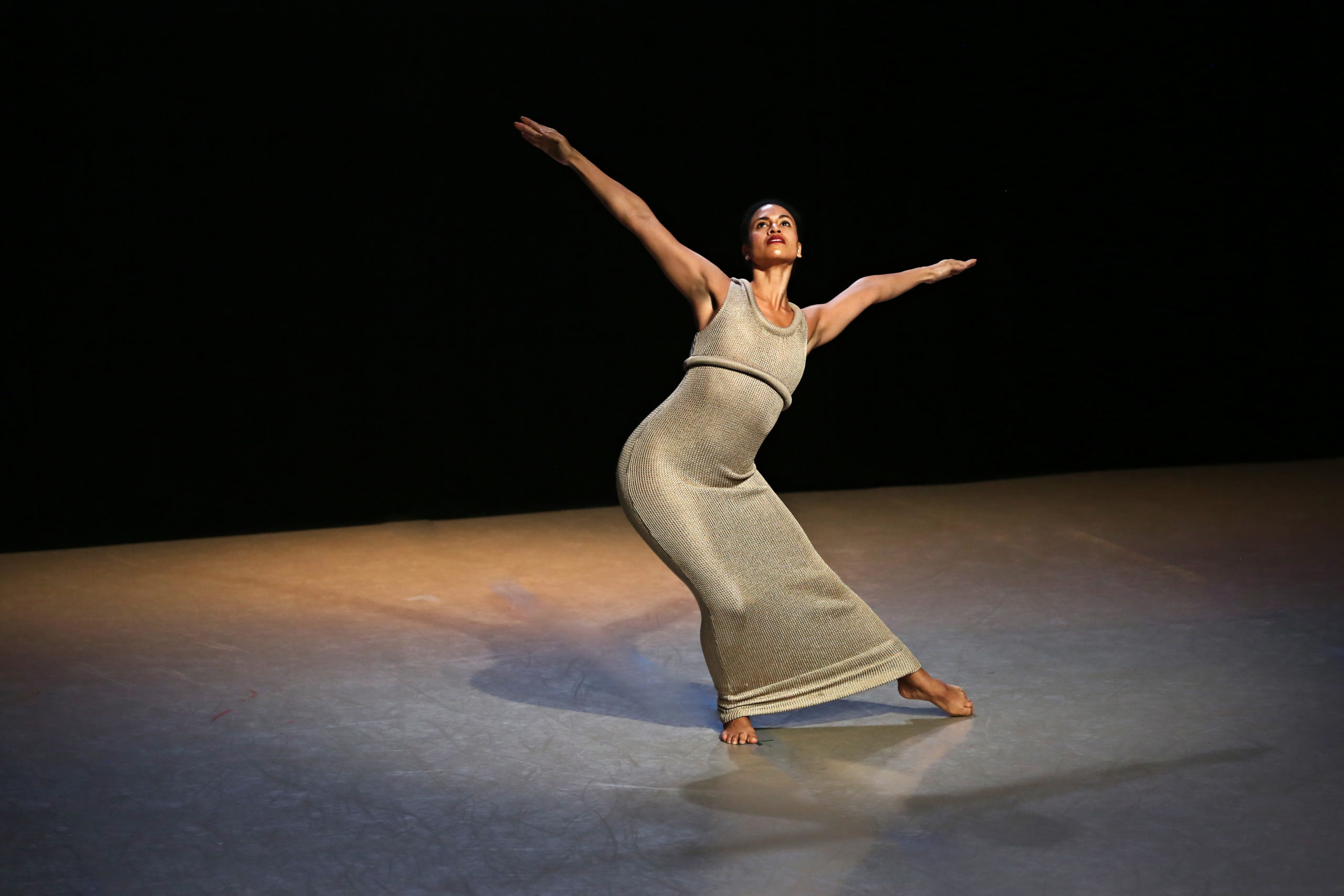 a dancer in a long grey dress stands on stage, knees bent and leaning into her right hip with arms splayed wide