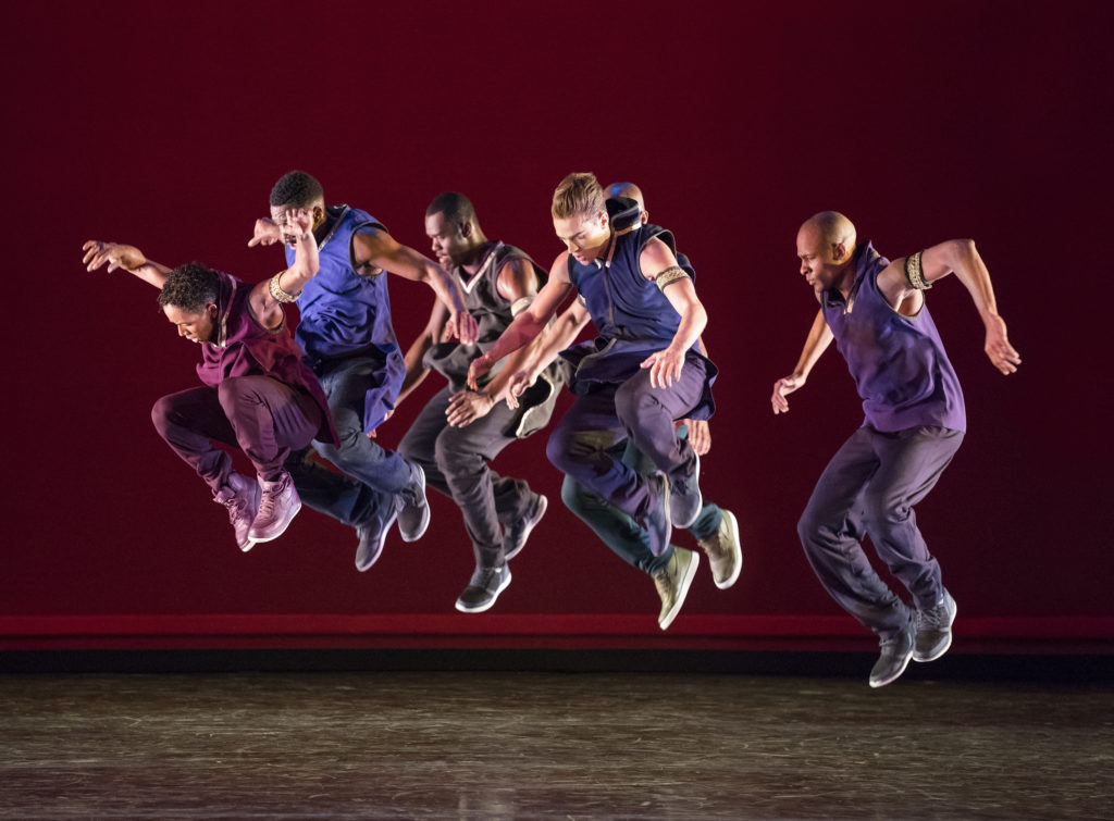 A group of six dancers dressed in shades of purple jump, lifting their knees and elbows, all facing the left of the frame.