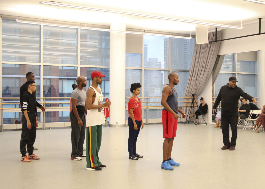 A group of seven dancers stand in a studio facing toward the right of the frame, where Rennie Harris stands, one arm to the side, demonstrating choreography.