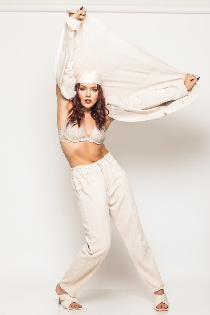 Cassidy Ratliff poses in a white outfit in front of a white background as she stretches an open white blouse behind her head.