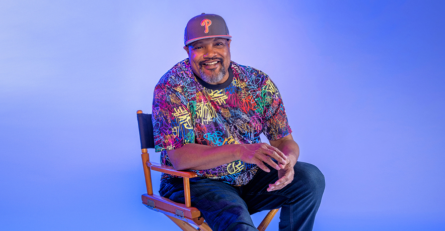 Rennie Harris sits in a director's chair in front of a purple background wearing a colorful t-shirt and jeans